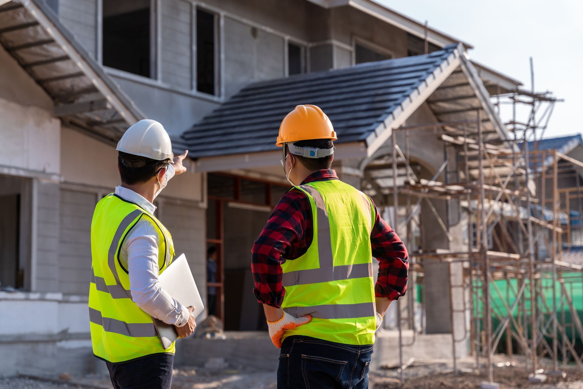 Deux ouvriers du bâtiment, vêtus de gilets et de casques de chantier, devant une maison en construction.