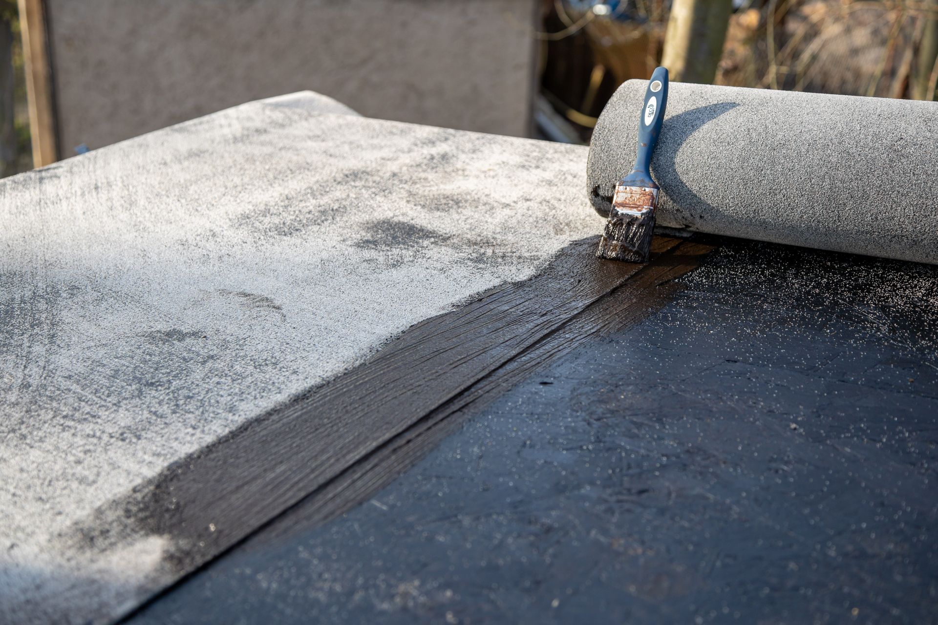 Application d'un mastic noir sur une surface de toiture grise et granuleuse à l'aide d'un pinceau et d'un rouleau.