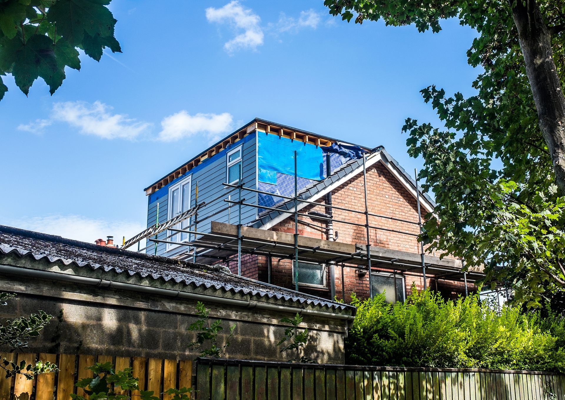 Maison en construction, façade en briques, échafaudages, bâche bleue recouvrant la partie supérieure, ciel ensoleillé.