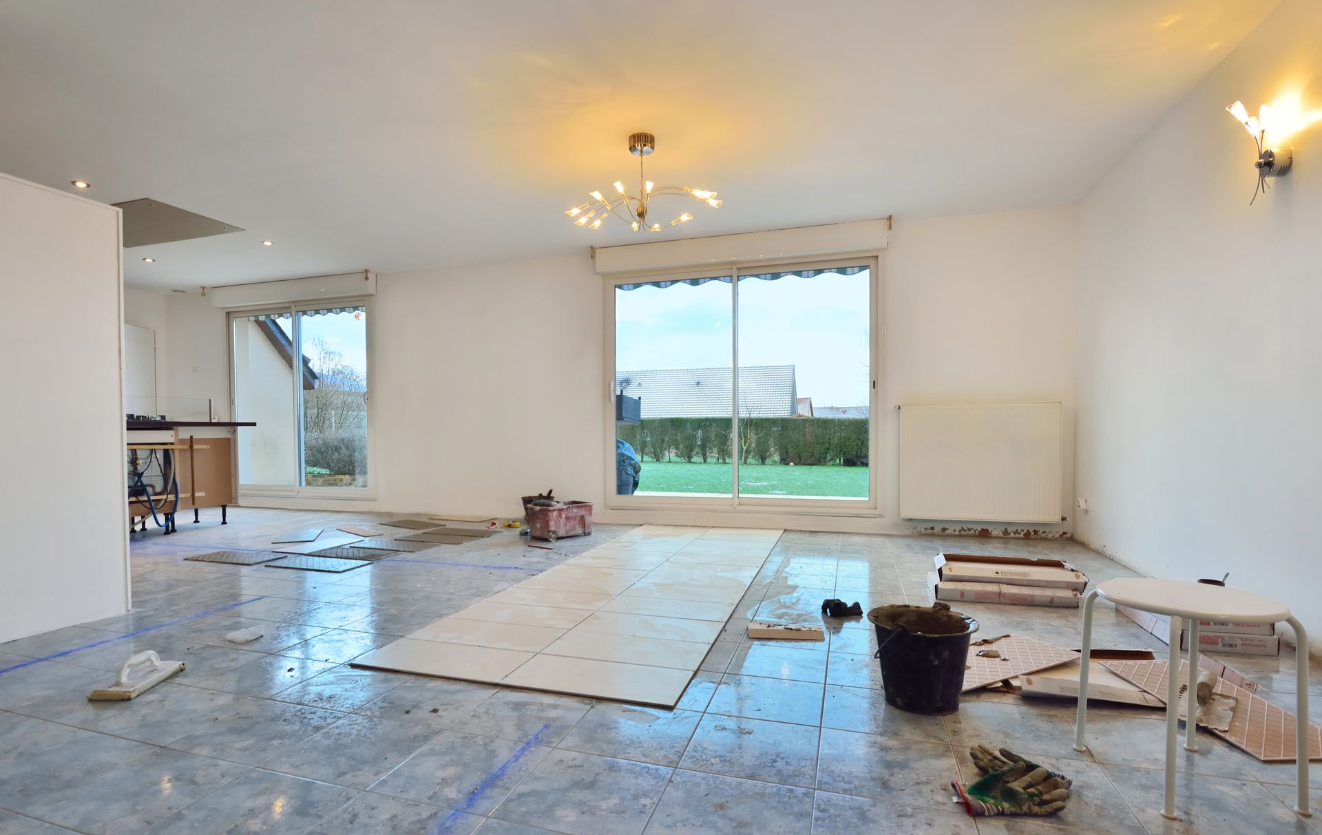 Intérieur en cours de rénovation ; espace ouvert avec de grandes fenêtres, sol non fini, outils et vue partielle sur l'extérieur.