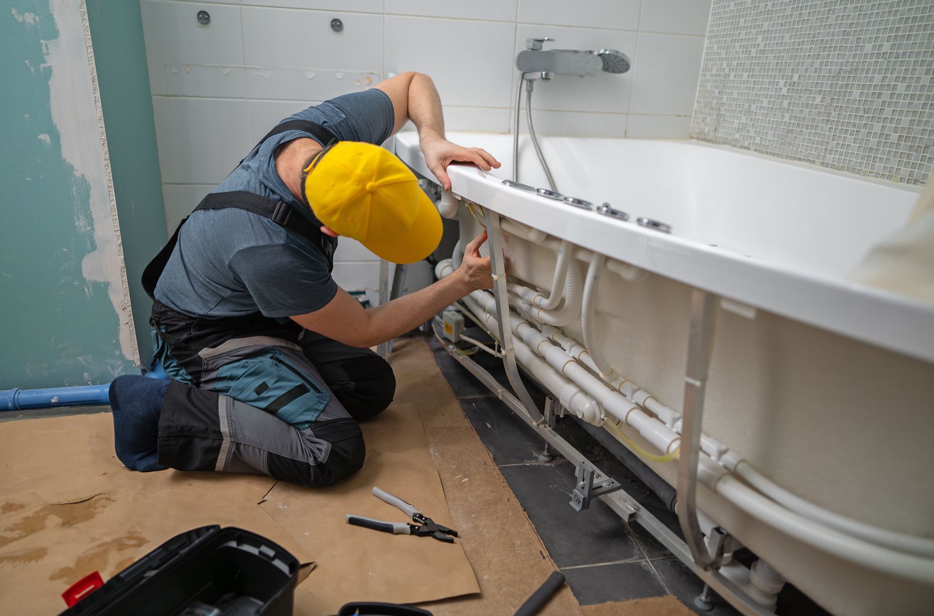 Ein Klempner mit gelbem Hut installiert während einer Renovierung die Rohrleitungen unter einer weißen Badewanne in einem Badezimmer.