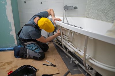 Ein Klempner mit gelbem Hut installiert während einer Renovierung die Rohrleitungen unter einer weißen Badewanne in einem Badezimmer.
