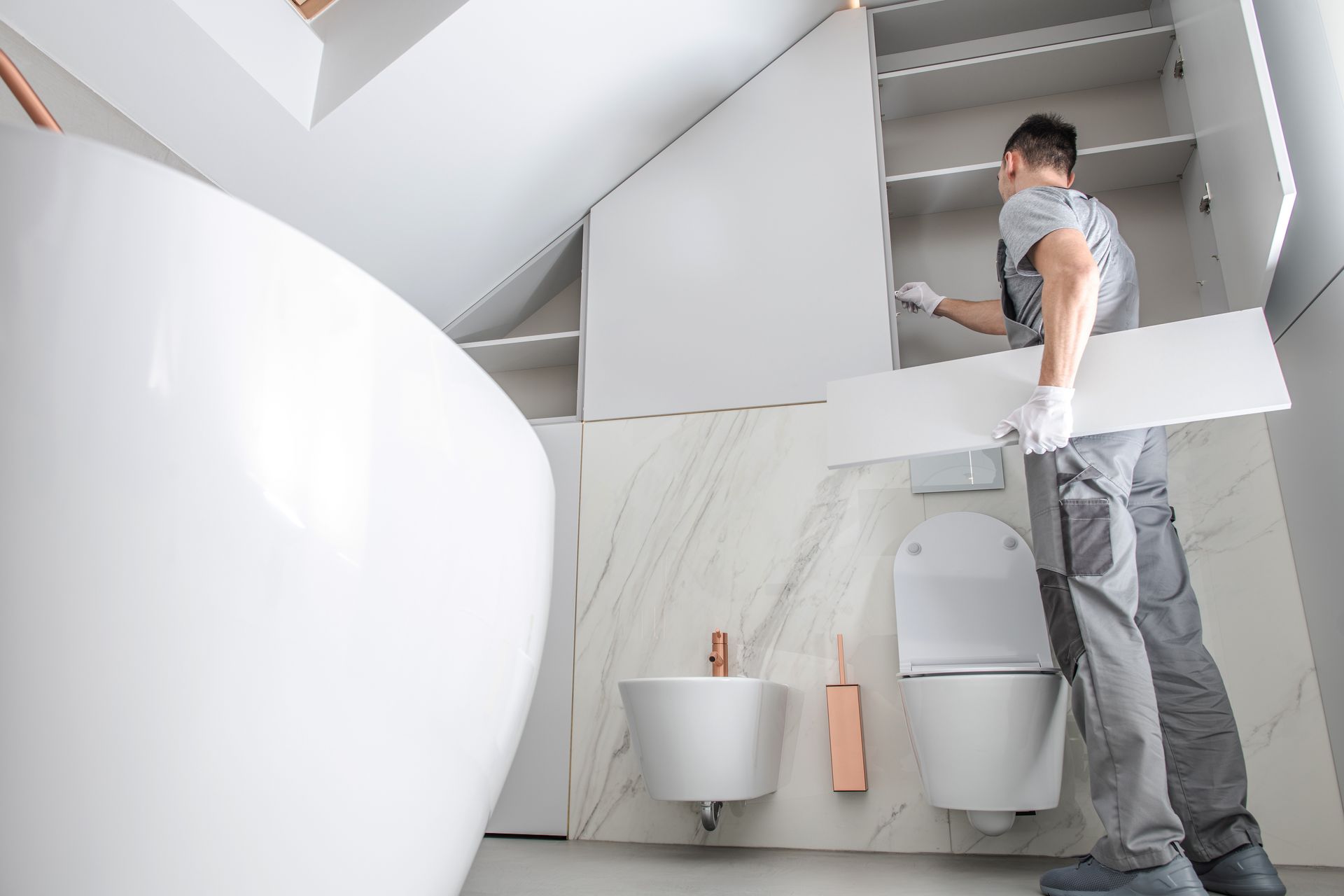 Mann installiert Regale in einem modernen weißen Badezimmer mit Toilette, Bidet und Badewanne.