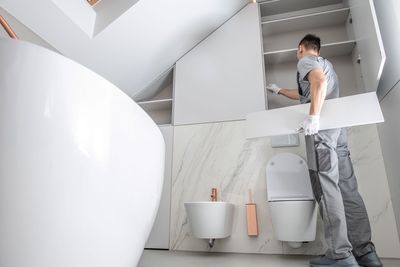 Mann installiert Regale in einem modernen weißen Badezimmer mit Toilette, Bidet und Badewanne.