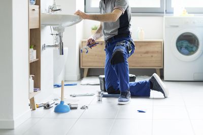 Klempner kniet neben Waschbecken und arbeitet mit Werkzeugen an der Wasserleitung in einem Badezimmer.