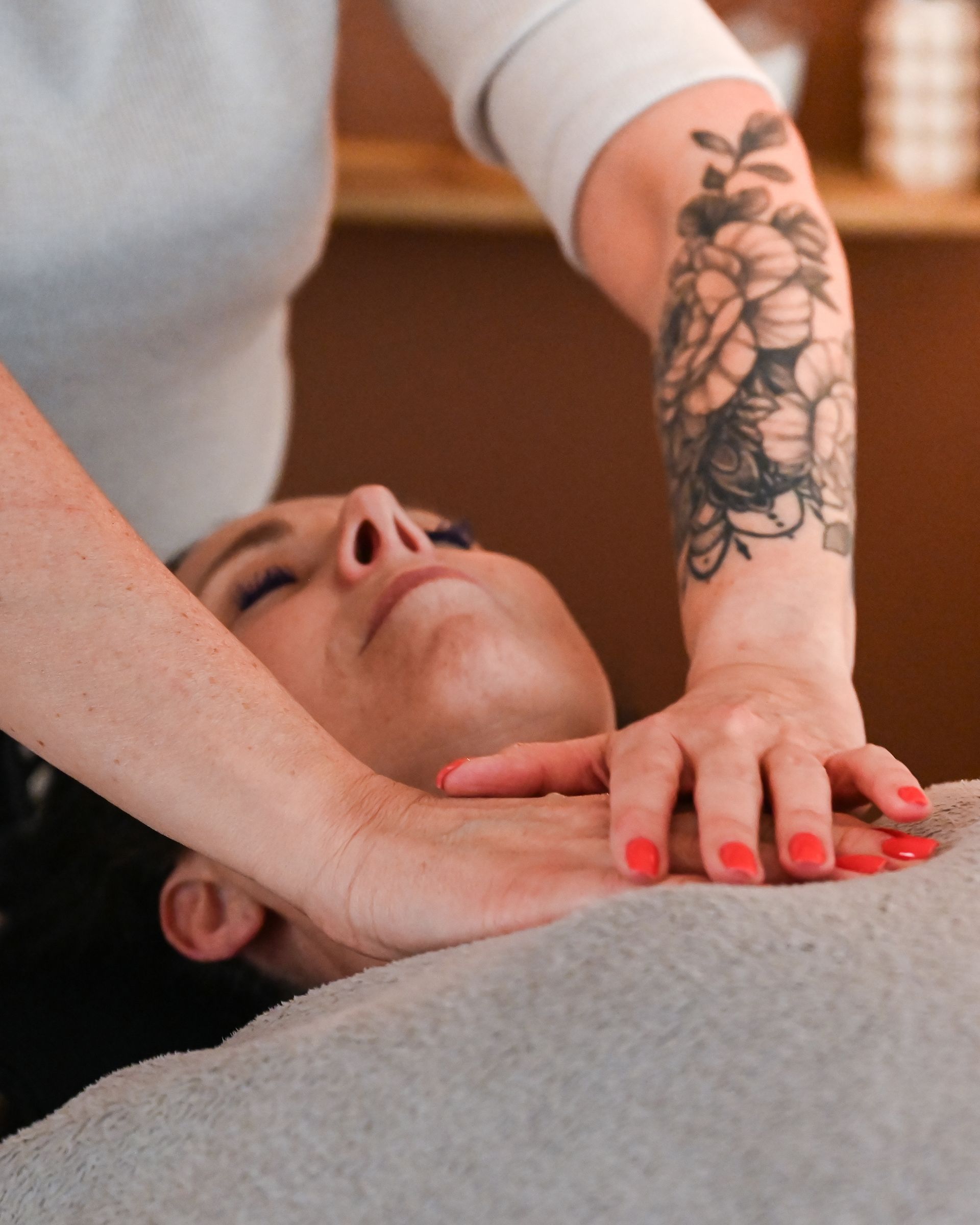Une masseuse effectue un massage de la nuque sur une cliente détendue, dont les ongles rouges et le tatouage floral sont visibles.