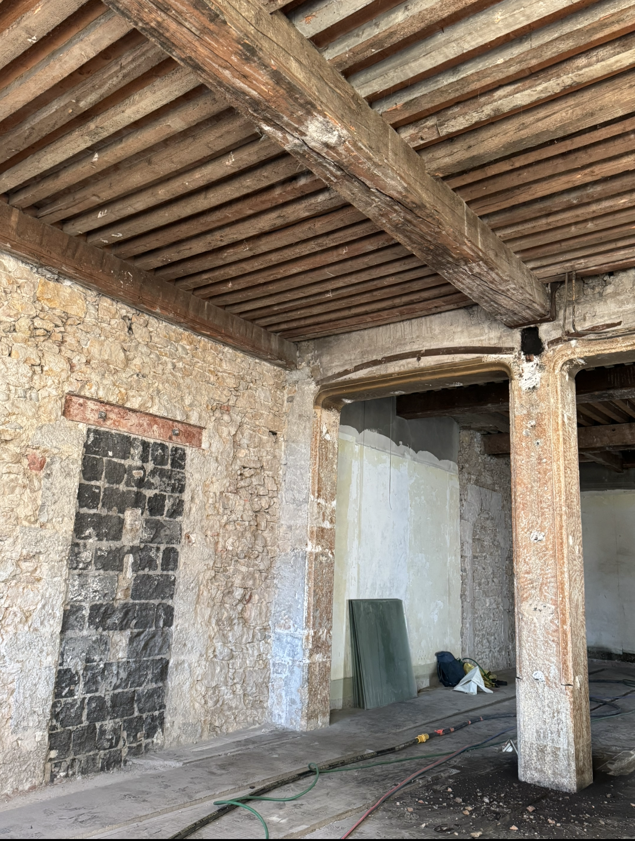 Vue intérieure d'un bâtiment avec des poutres en bois apparentes, des murs en pierre et une porte condamnée.