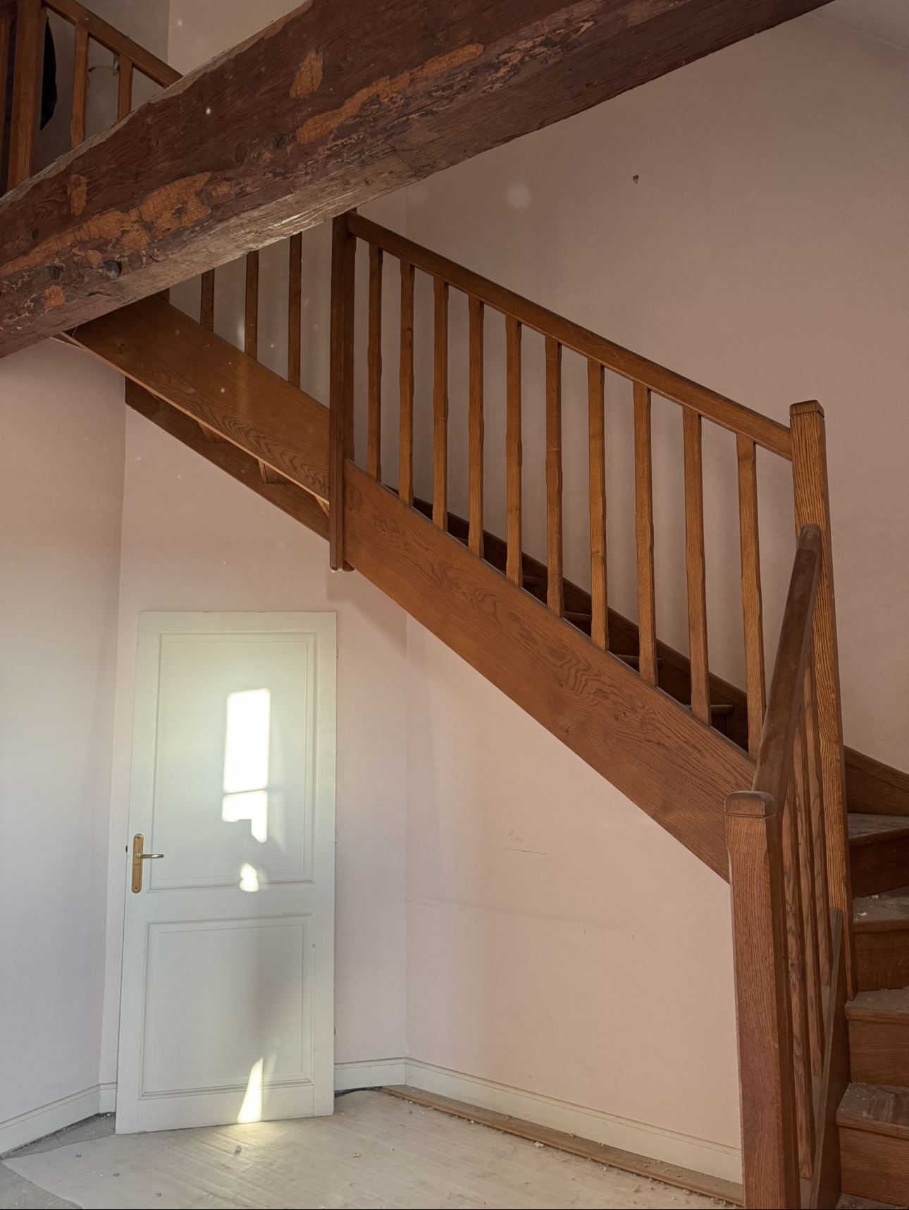 Escalier en bois avec rampe, porte et poutres apparentes, dans une pièce aux murs rose pâle.