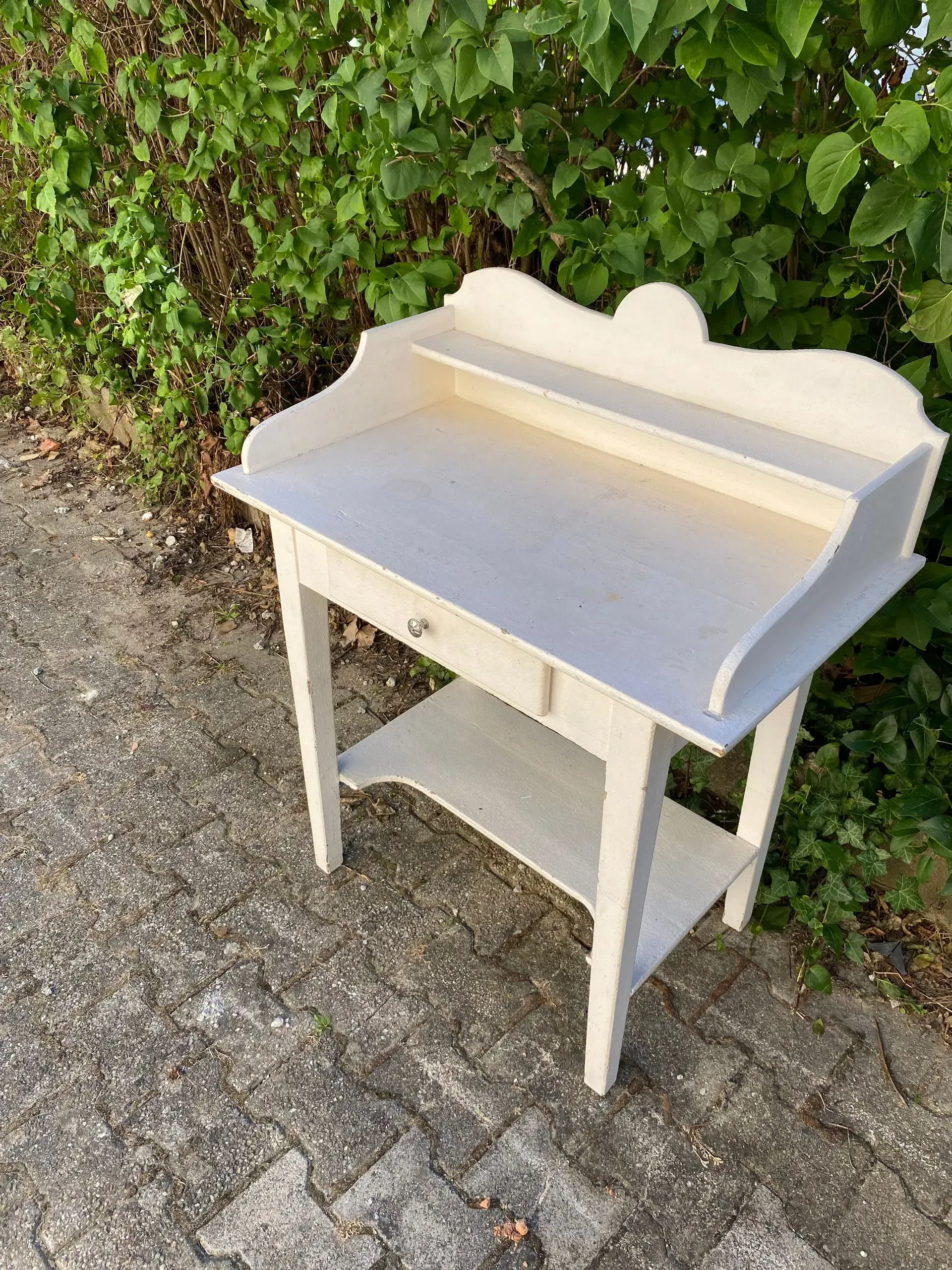 Table d'appoint en bois blanc avec tiroir, étagère et plateau décoratif, posée à l'extérieur.