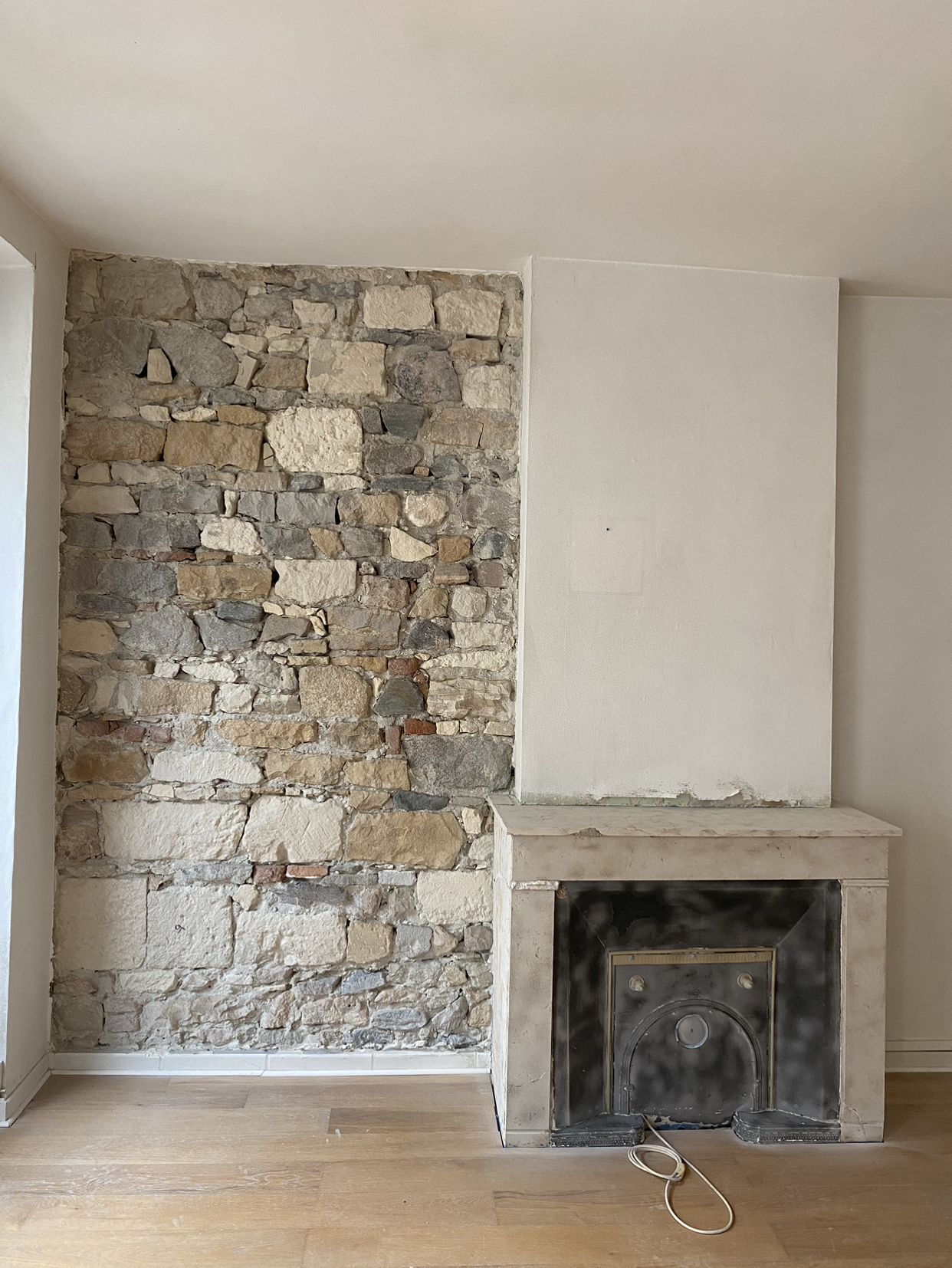 Mur en pierre et cheminée, intérieur inachevé. Sol en bois, enduit et murs couleur crème.