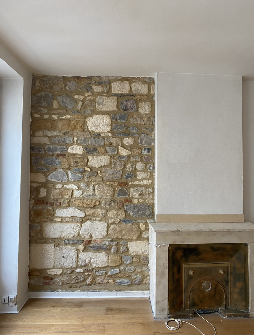 Fond en pierre avec cheminée et pan de mur peint en blanc. Sol en bois.