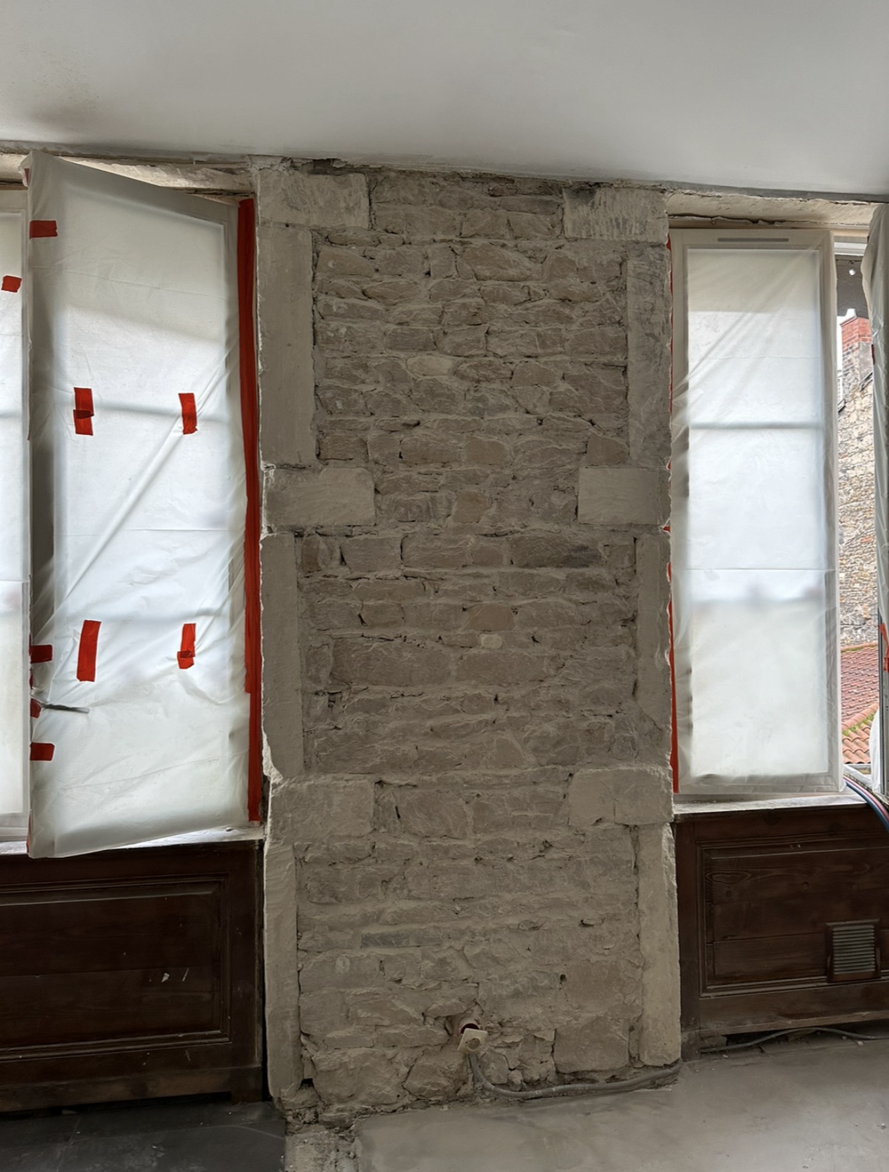 Intérieur d'une pièce aux murs de pierre, fenêtres recouvertes de plastique et briques apparentes.