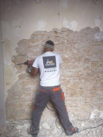 Personne en tenue de travail utilisant une perceuse sur un mur pour enlever du plâtre.