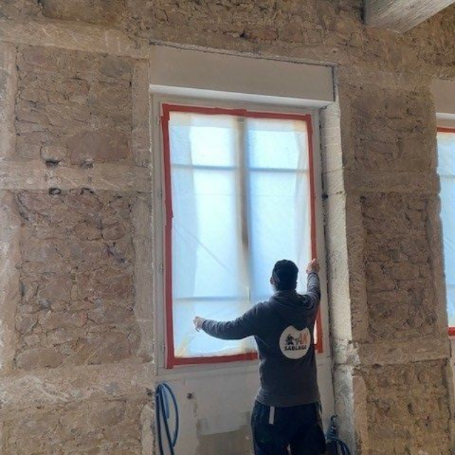 Un homme fixe une bâche en plastique sur le cadre d'une fenêtre dans une pièce aux murs en pierre.