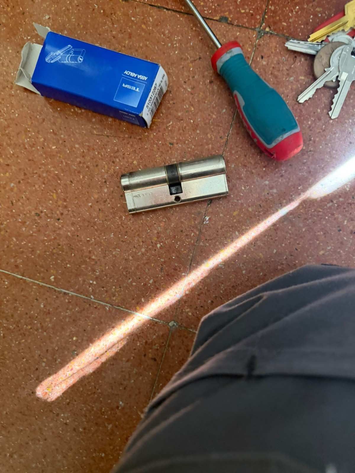 Herramientas sobre un suelo de baldosas: caja azul, destornillador, cilindro de cerradura y llaves, con la rodilla de una persona visible.