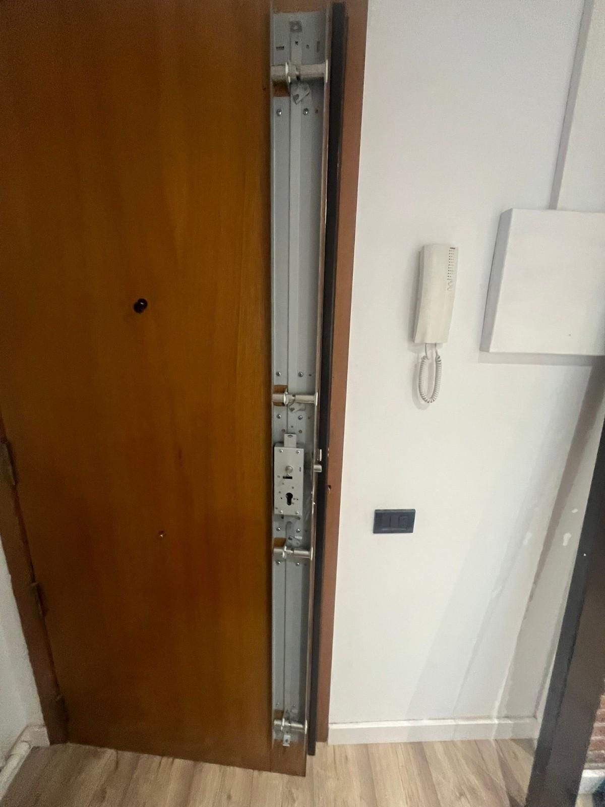 Puerta de madera en un apartamento con un estrecho mecanismo de cerradura de metal a la vista, junto a una pared blanca y un interfono.