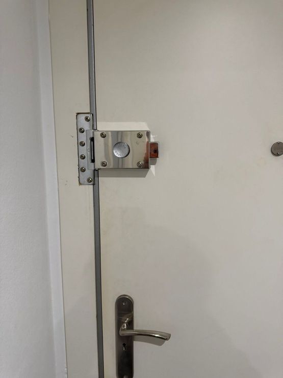 Primer plano de una puerta blanca con pestillo, manija y bisagra de metal en una pared lisa.
