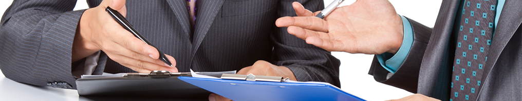 Dos personas vestidas con traje de negocios están sentadas a una mesa con una carpeta azul; una gesticula mientras la otra sostiene un bolígrafo.