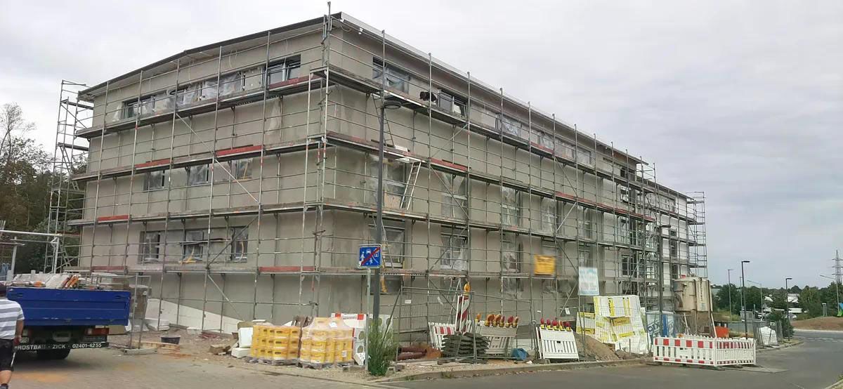 Gebäude im Bau mit Gerüst, umgeben von Baumaterialien, an einem bewölkten Tag. Ein blauer LKW ist teilweise sichtbar.