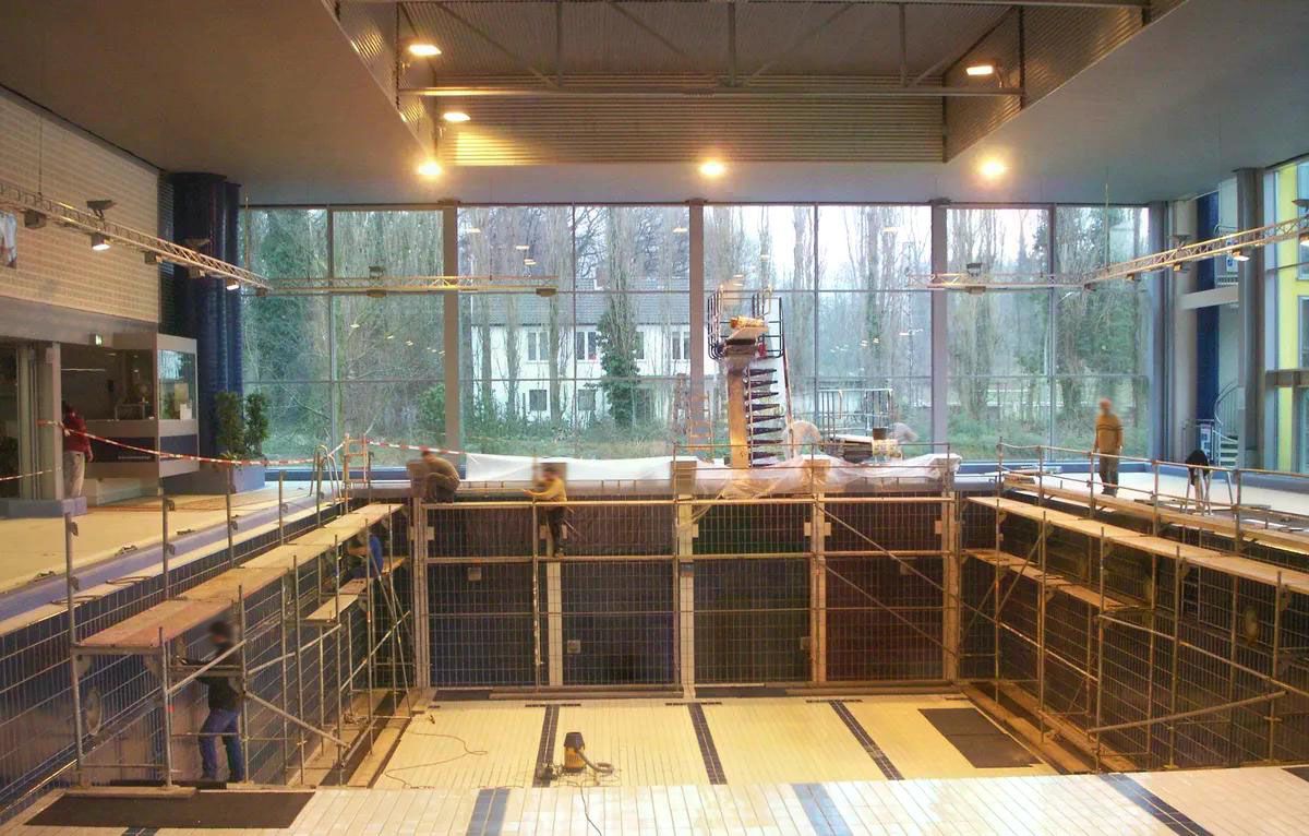 Leeres Hallenbad, das gerade renoviert wird. Die Wände sind mit Gerüsten verkleidet und durch ein großes Fenster blicken Sie auf Bäume.