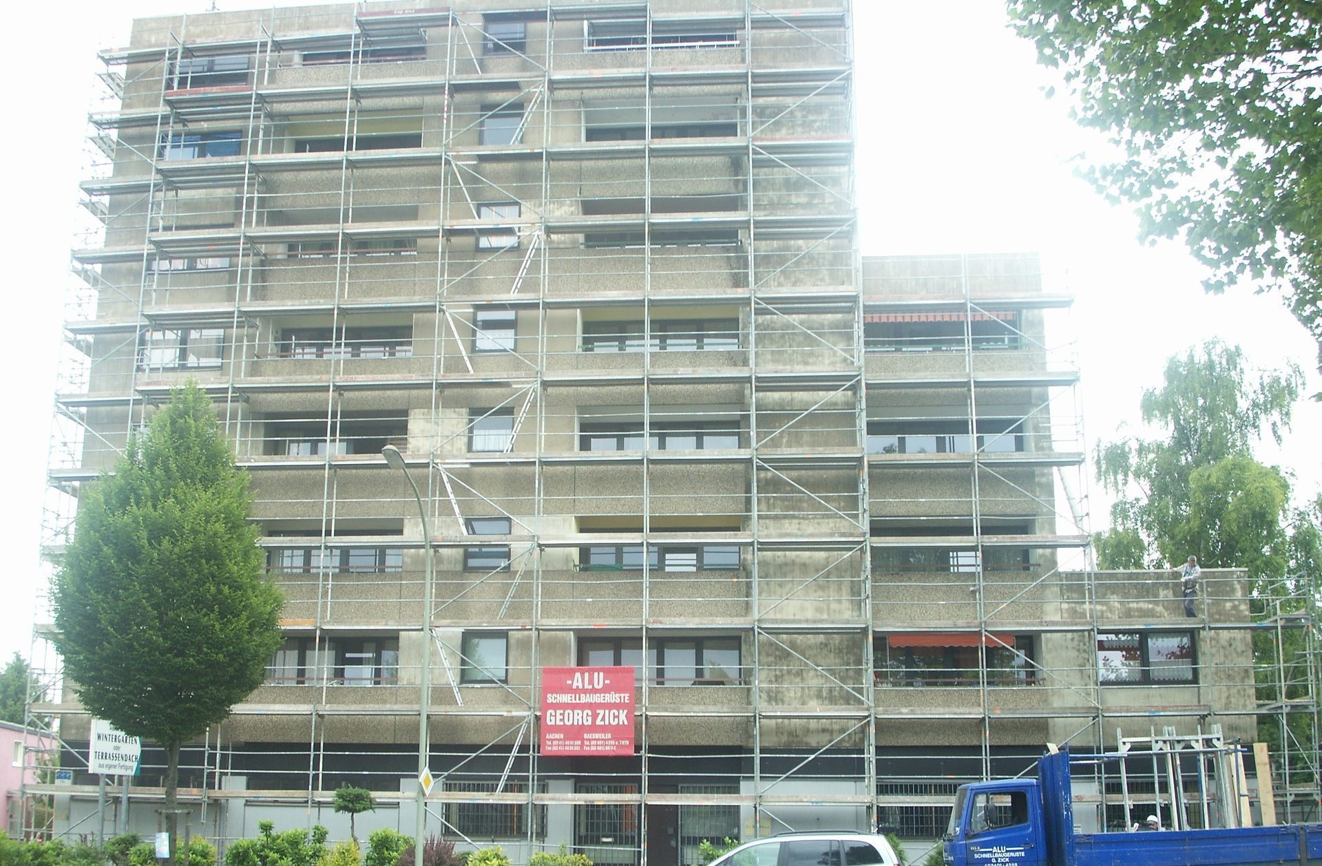 Ein mehrstöckiges Gebäude im Bau, eingerüstet. Im Vordergrund steht ein kleiner Baum, davor ein blauer Lastwagen.