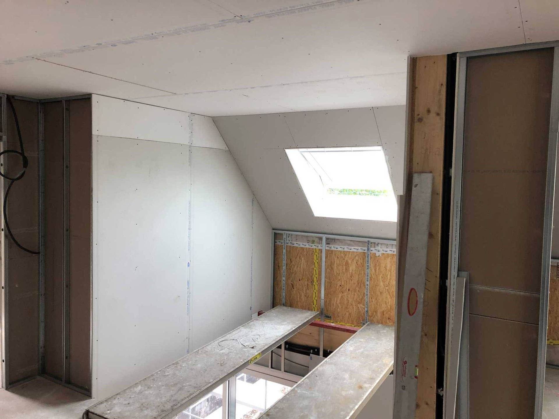 Combles en cours d’aménagement, avec des plaques de plâtre, une charpente apparente et un puits de lumière au plafond