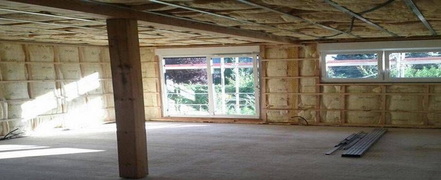 Intérieur inachevé avec une ossature apparente, un poteau de soutien, de grandes fenêtres et la lumière du soleil sur le sol.