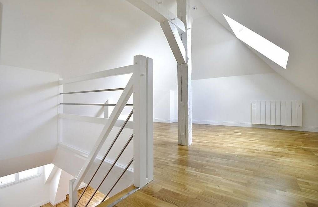 Pièce mansardée lumineuse avec parquet, fenêtre de toit, murs blancs et rampe d’escalier