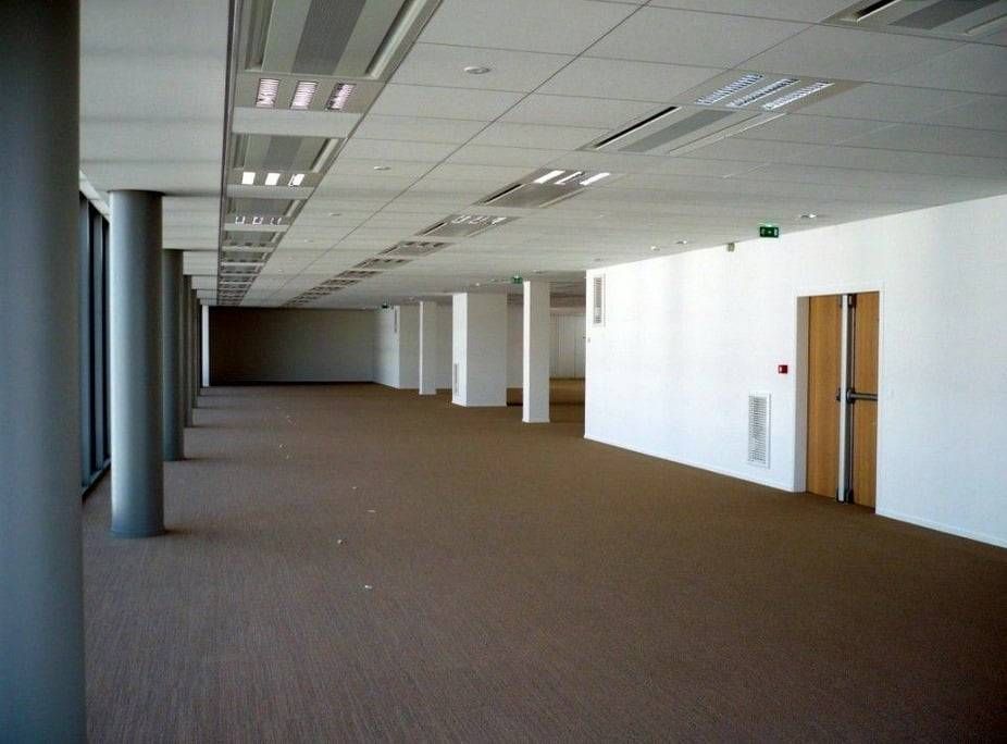 Couloir de bureaux vide avec de la moquette, des murs blancs et de grandes fenêtres sur la gauche
