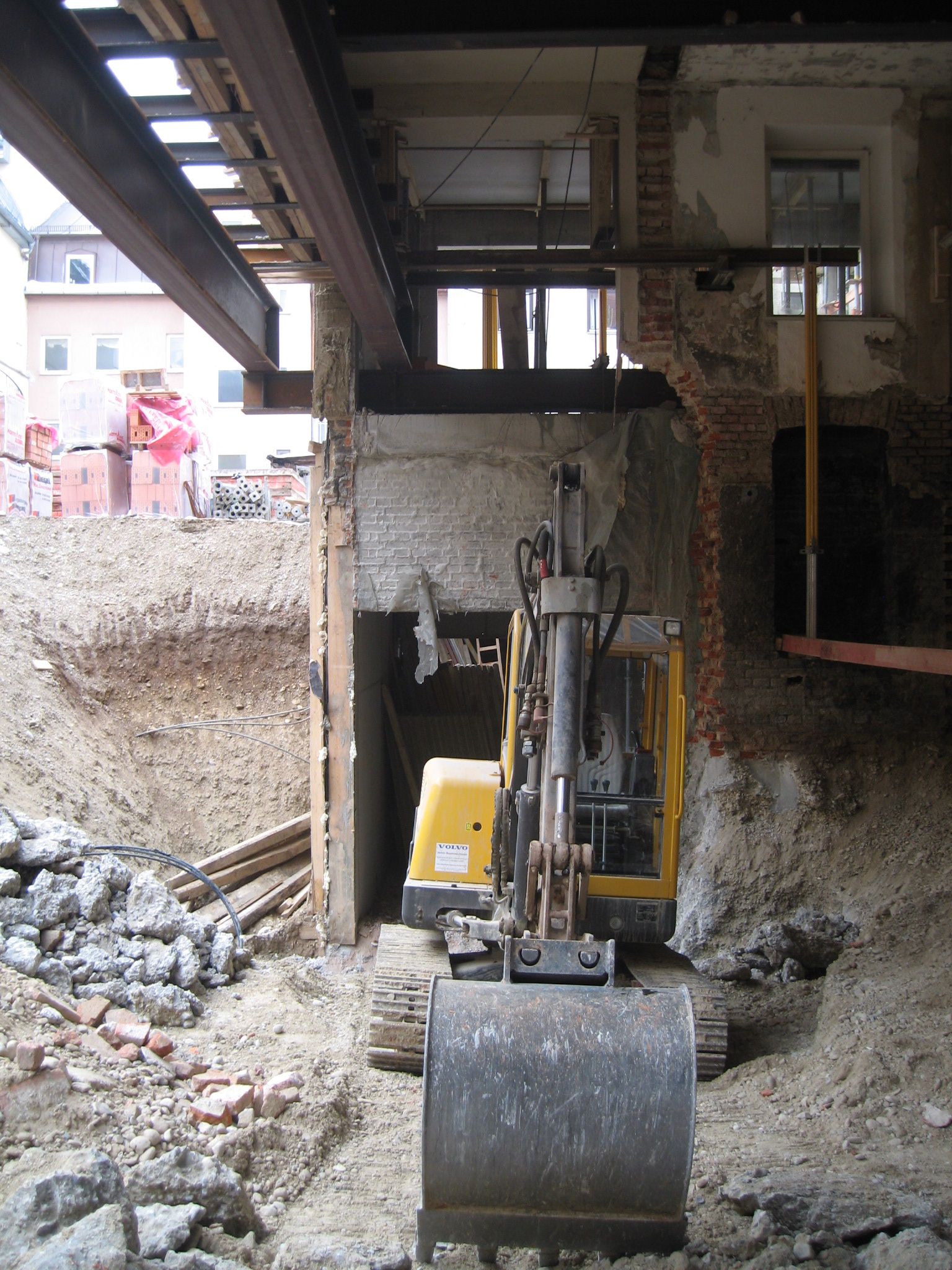 Ein gelber Bagger bei präzisen Rückbauarbeiten in einer schmalen Baugrube unter einem Gebäude.