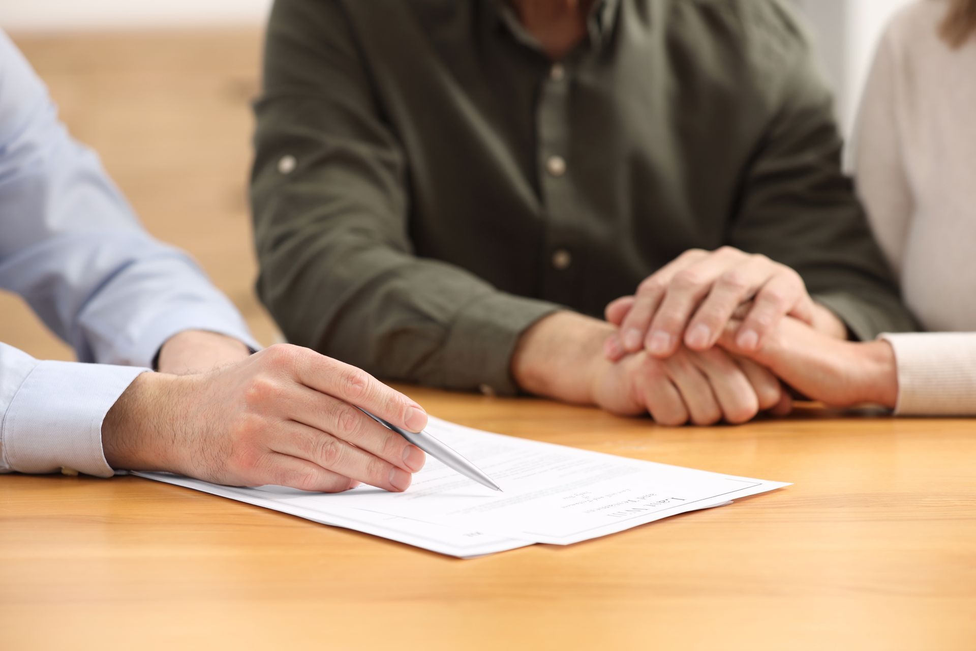 Une personne écrit sur un document à une table en bois, tandis qu’une autre pose ses mains jointes à côté d’elle.