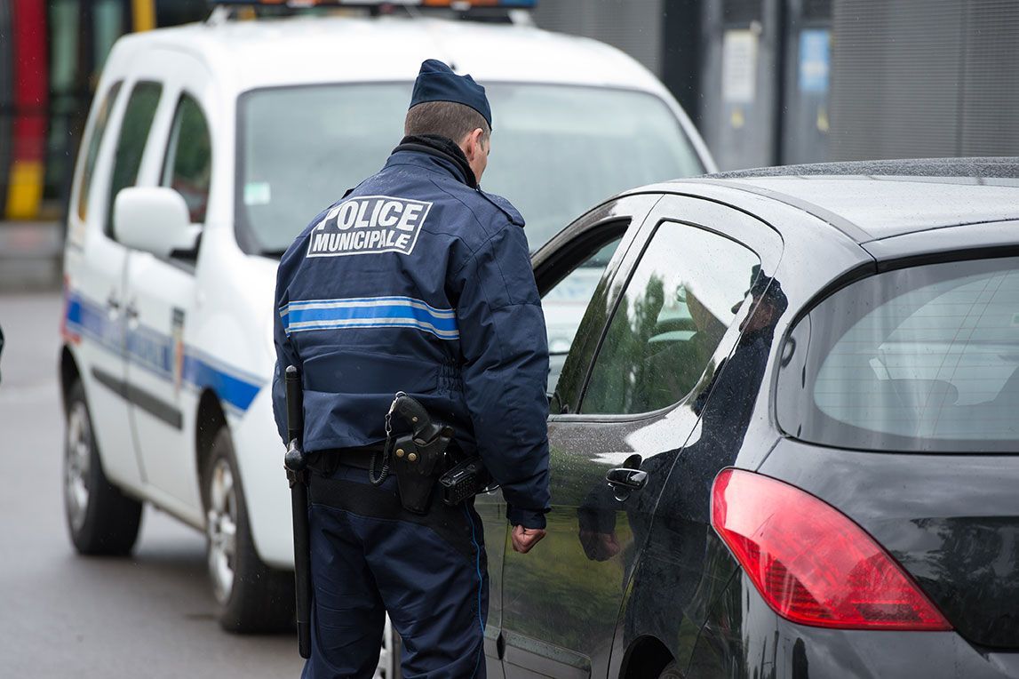Un policier inspecte une voiture, avec un véhicule de patrouille en arrière-plan.
