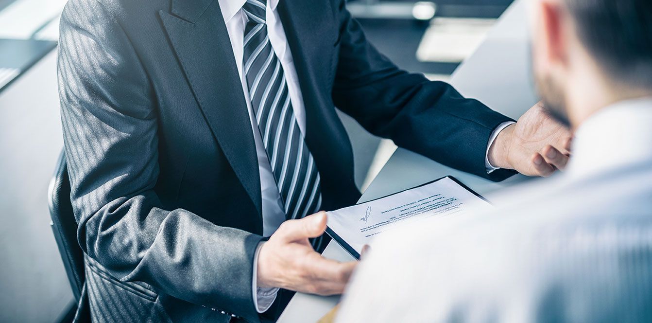 Un homme en costume montre du doigt un document qu'il tient tout en parlant à un autre homme.