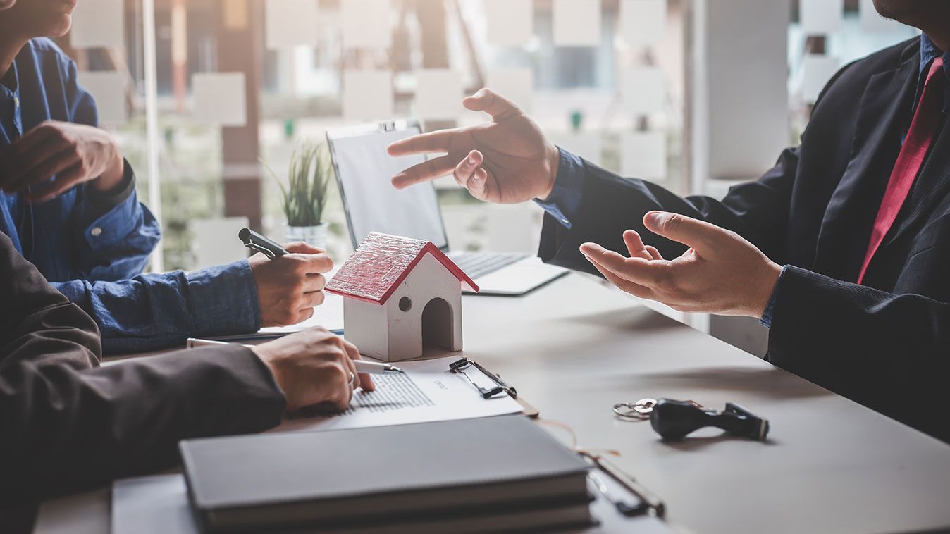 Des gens en costume discutent de paperasse autour d'une table avec une maison miniature, des clés et un ordinateur portable.