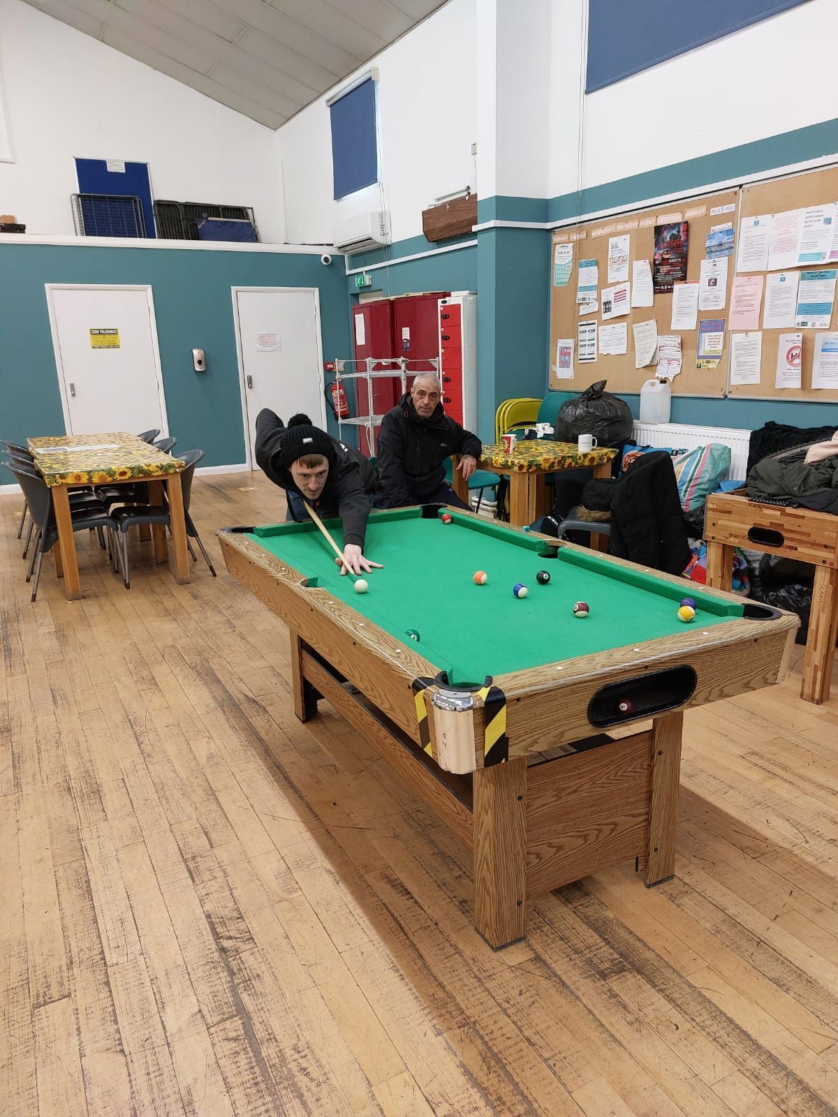 A man is playing pool in a room with tables and chairs.