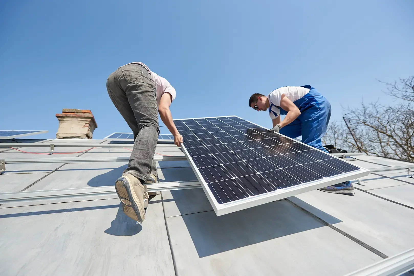 Zwei Arbeiter installieren vor strahlend blauem Himmel ein Solarpanel auf einem Dach.