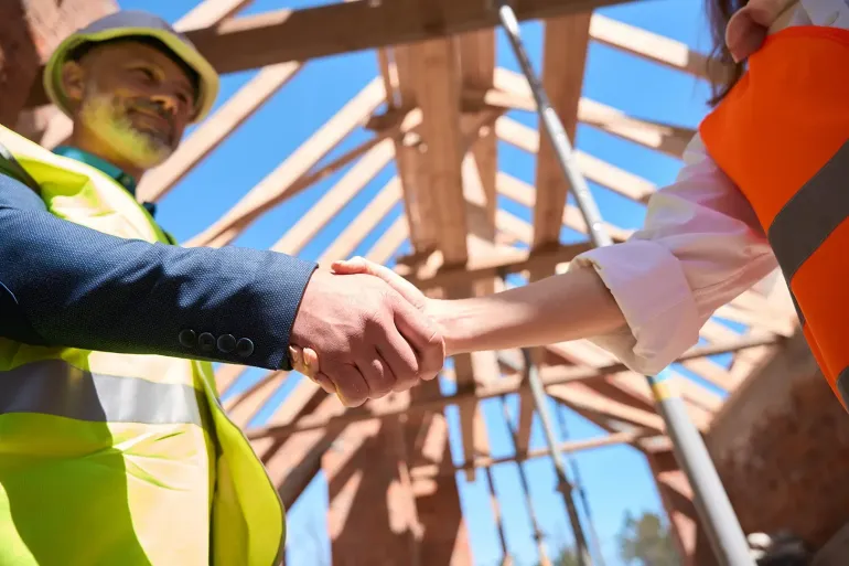 Zwei Personen in Sicherheitswesten schütteln sich auf einer Baustelle die Hände. Holzbalken und blauer Himmel sind zu sehen.