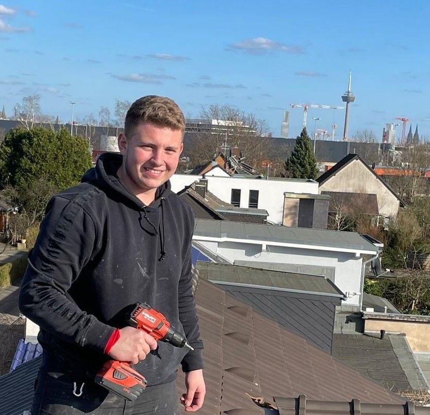 Zwei Dachdecker mit Schutzhelmen und Warnwesten geben sich auf einem frisch gedeckten Ziegeldach ein High Five; im Hintergrund ist der Sonnenuntergang zu sehen.