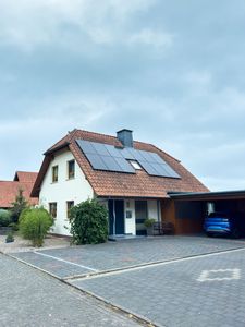 Haus mit Solarzellen auf dem Dach, ein blaues Auto im Carport und ein bewölkter Himmel.