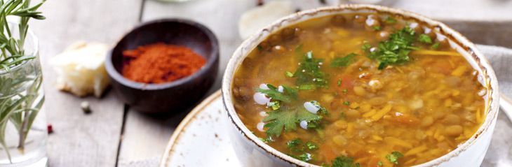 Un tazón de sopa de lentejas adornado con hierbas junto a un pequeño plato de especias rojas en una mesa rústica.