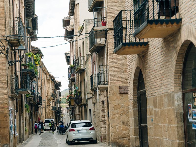 Callejón estrecho de piedra con balcones y coches aparcados en un pueblo histórico.