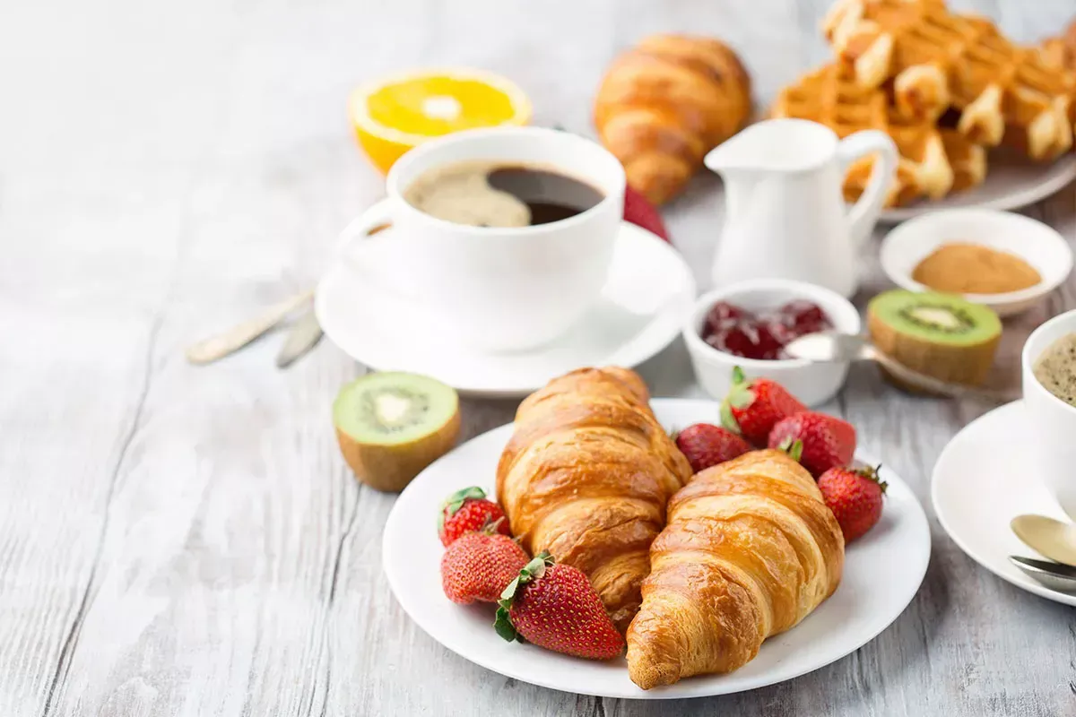 Croissants, fresas, kiwi y café en una mesa de desayuno luminosa.