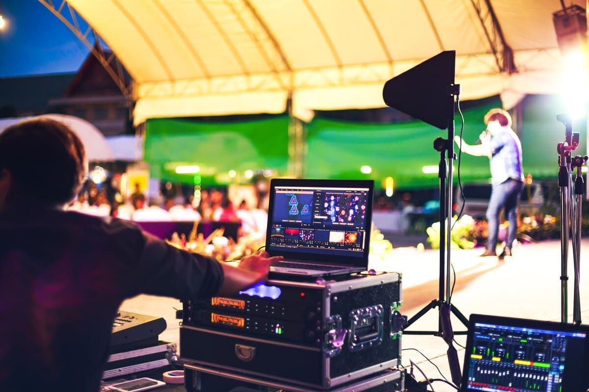 Una persona operando equipos de sonido y video en un evento al aire libre. Un artista está en el escenario frente al público.