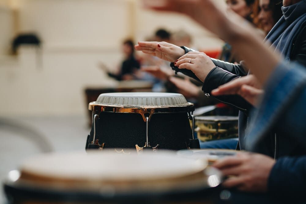 Personas tocando la batería en una clase, con las manos levantadas. Tambores negros con aros dorados; fondo borroso.