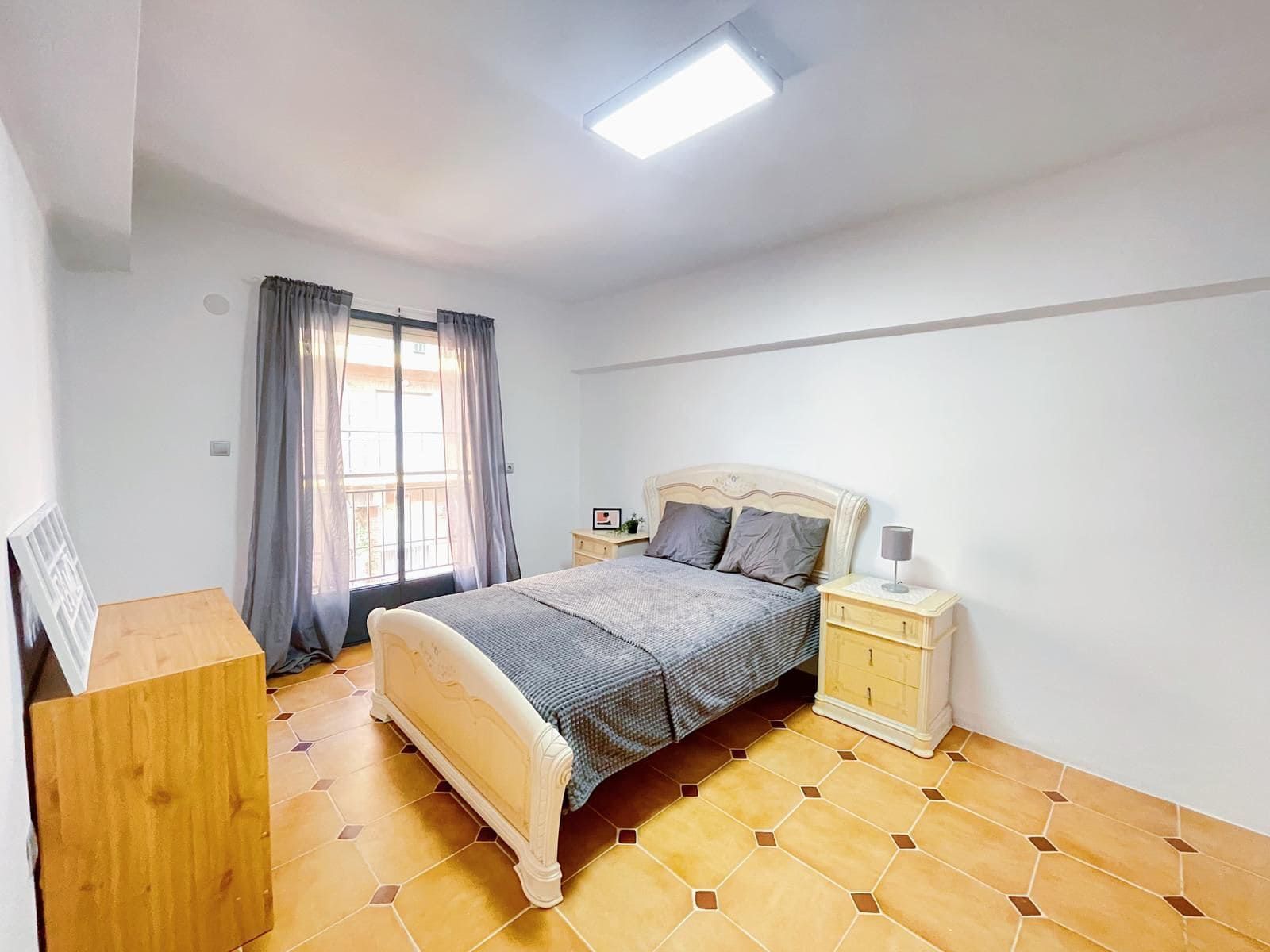 Dormitorio con cama, mesitas de noche, cómoda y ventana con cortinas. Suelo de madera.