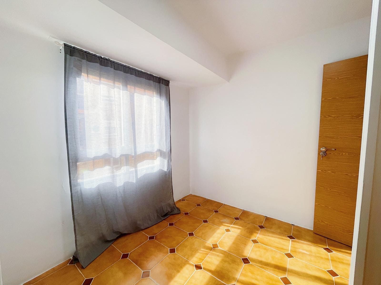 Habitación pequeña y vacía con suelo de baldosas, una ventana con cortinas grises y una puerta de madera.