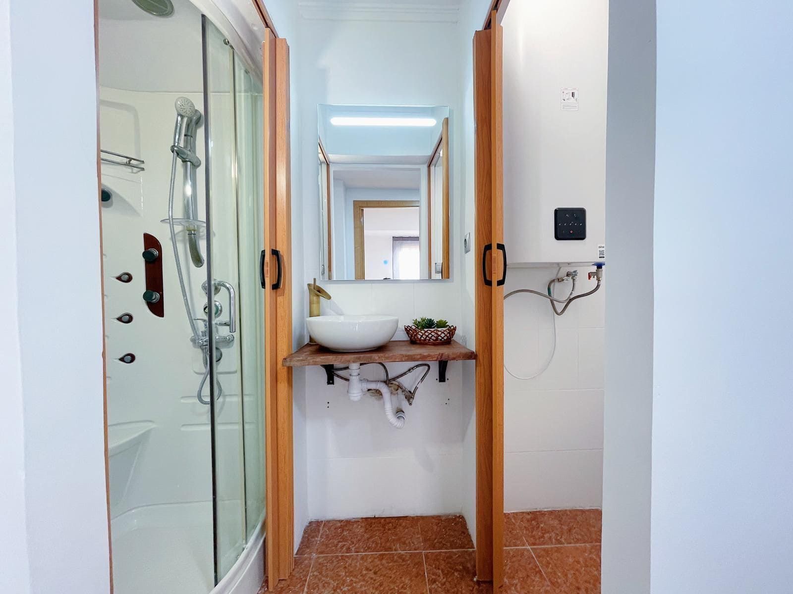 Baño pequeño con ducha, lavabo y calentador de agua. Puertas y grifería marrones, paredes blancas y suelo marrón rojizo.