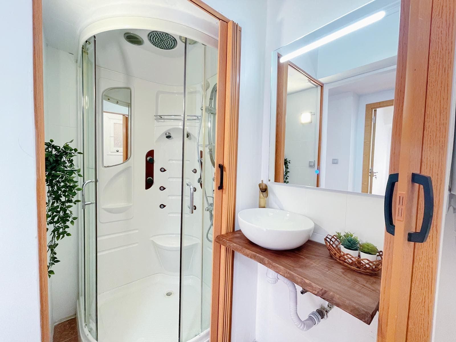 Baño con ducha de cristal, detalles de madera y lavabo blanco sobre estante de madera.