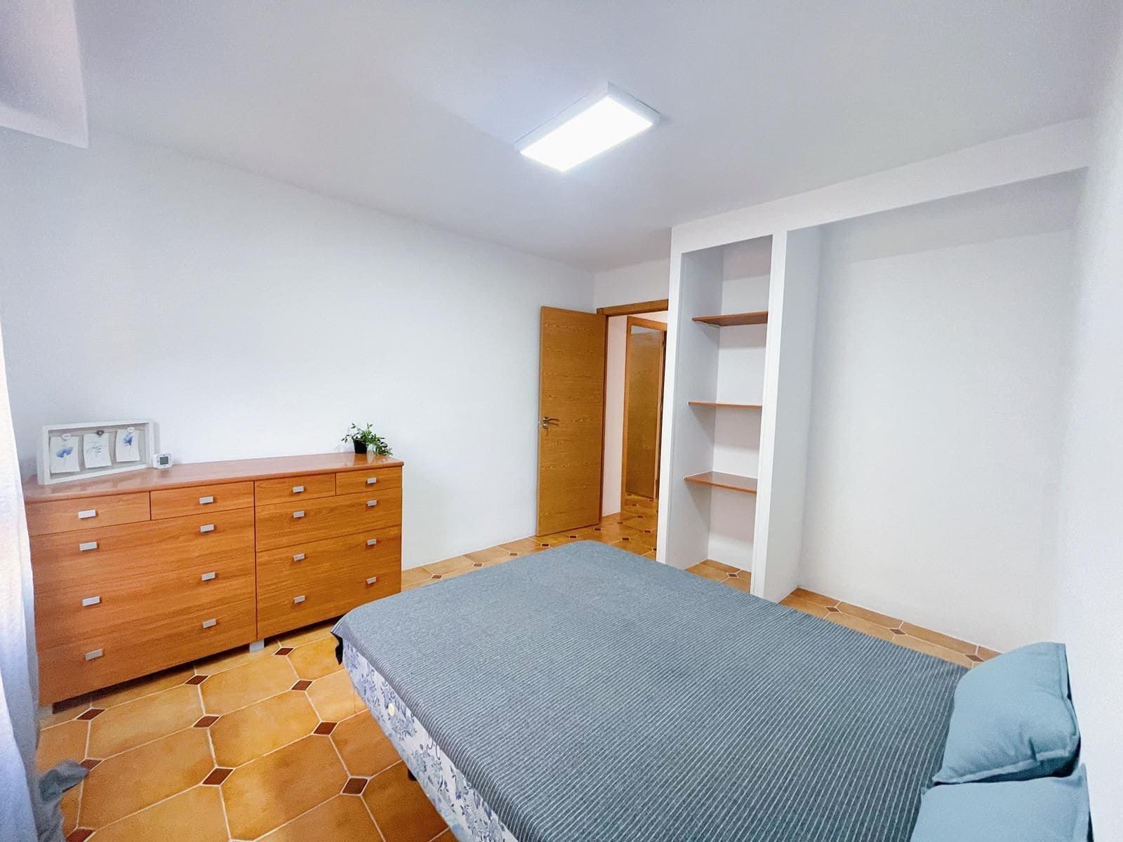 Dormitorio con cómoda de madera, cama, estantería y puerta abierta. Paredes claras y suelo de madera.