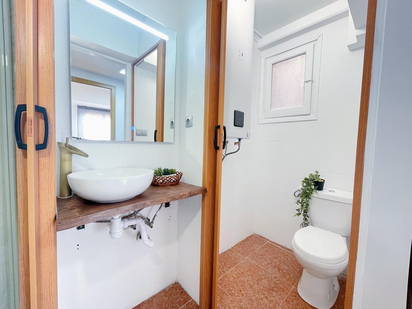 Pequeño baño con lavabo blanco, tocador de madera e inodoro; tonos de madera cálidos y paredes blancas.
